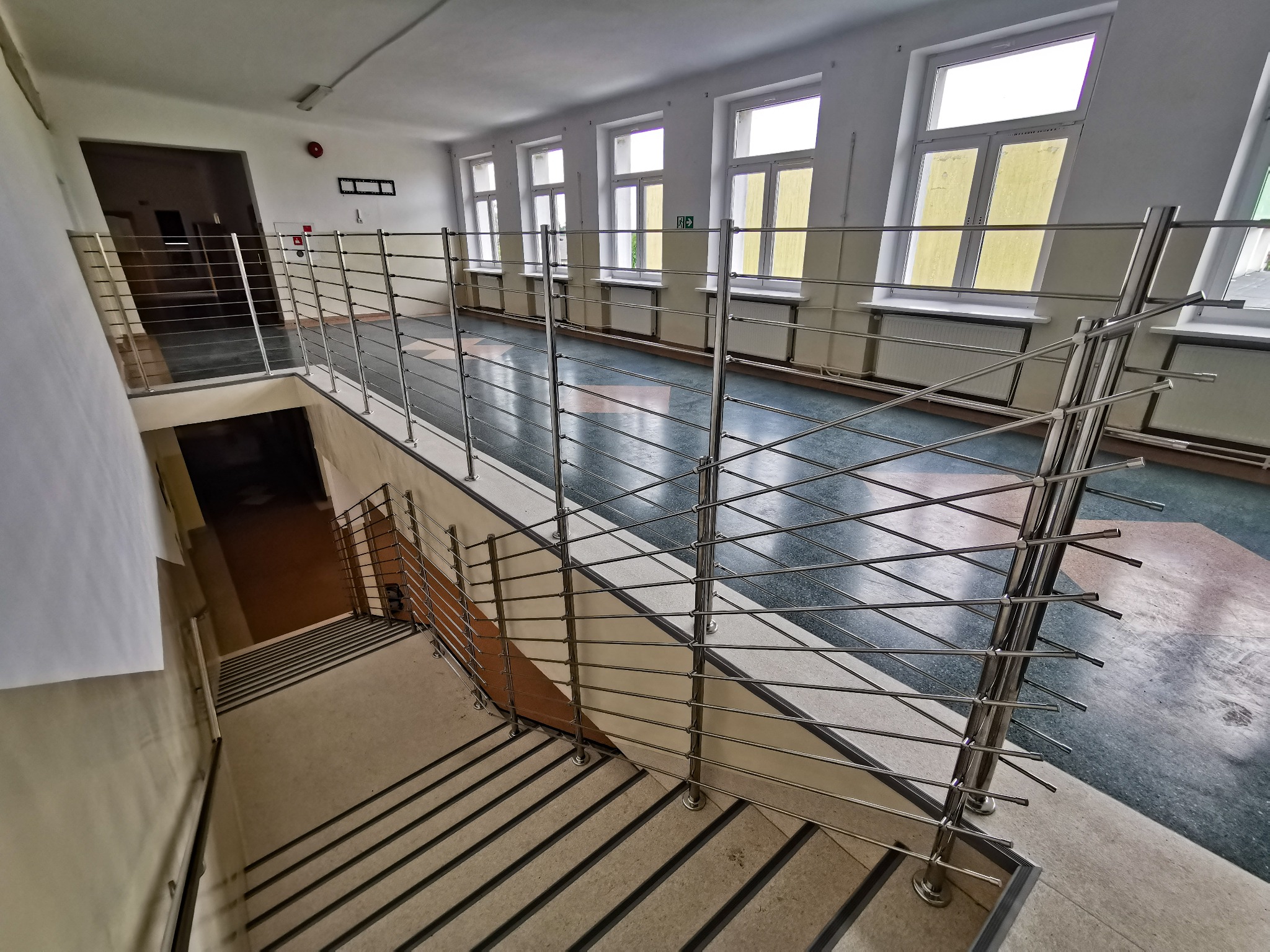 Wnętrze budynku z widokiem na schody i poziom wyżej, zabezpieczone metalową balustradą z poziomych rurek, duże okna w tle.