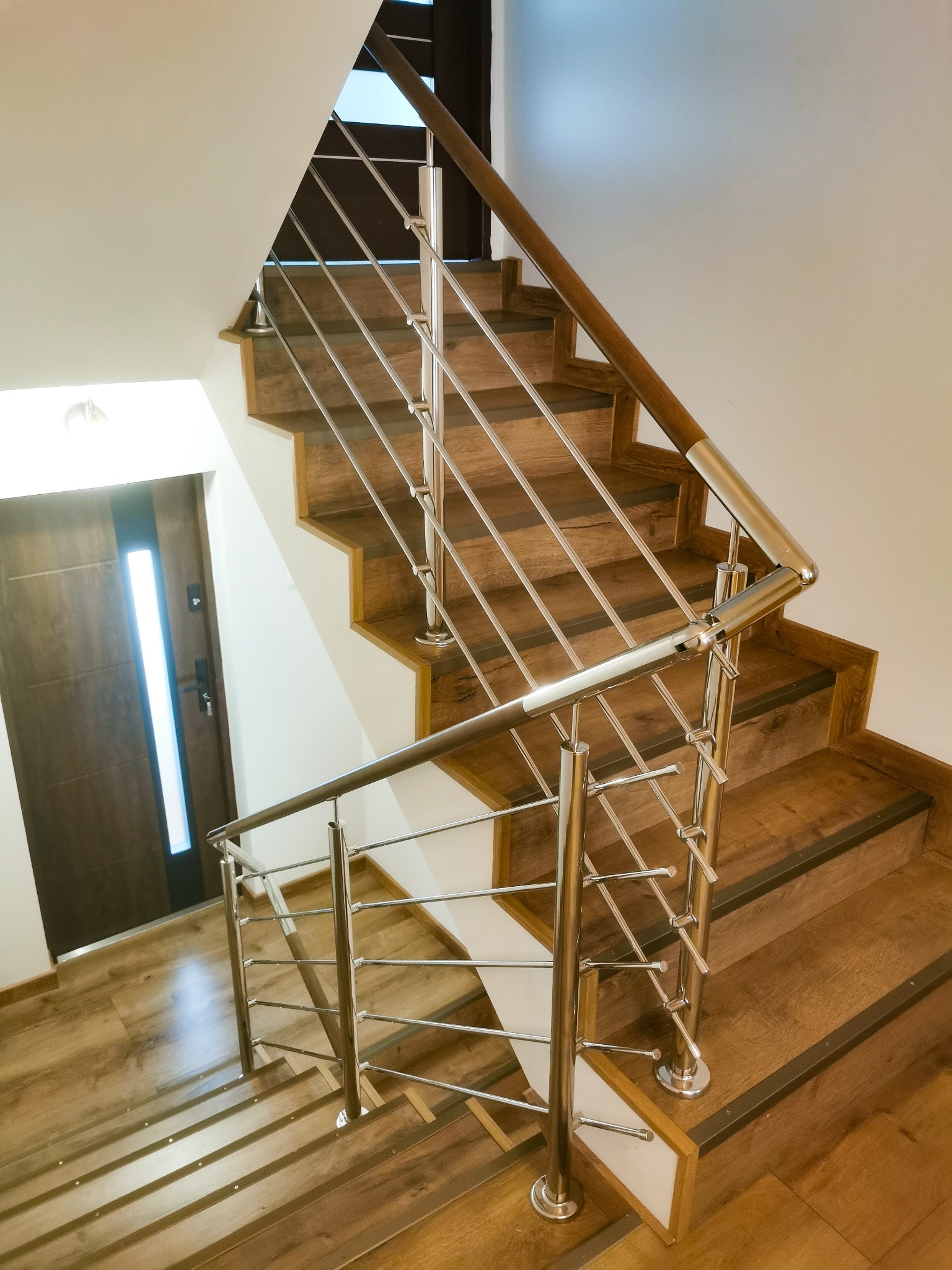 Schody drewniane z polerowaną, metalową balustradą z poziomymi prętami, widok z góry, brązowe drzwi wejściowe w tle.