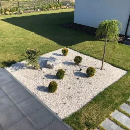 Nowoczesny ogród z widokiem z góry: geometryczny wzór z białego żwiru, krzewów, pojedynczego kamienia i drzewa płaczącego, oświetlony małymi lampami, obok trawnik i ścieżka z płyt.