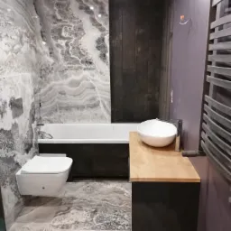 Nowoczesna łazienka z marmurowymi płytkami na ścianach i podłodze, wisząca toaleta i umywalka na drewnianym blacie, grzejnik drabinkowy oraz ciemne panele ścienne, widoczne niedokończone...