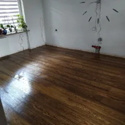 Pokój po cyklinowaniu i lakierowaniu parkietu, widoczne gniazdka elektryczne bez osprzętu, zegar ścienny z wystającymi przewodami, parapet z roślinami.