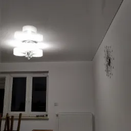 Biały, błyszczący sufit napinany z zamontowaną, nowoczesną lampą z kryształkami, widoczny zegar słoneczny na ścianie, okno i grzejnik w tle.