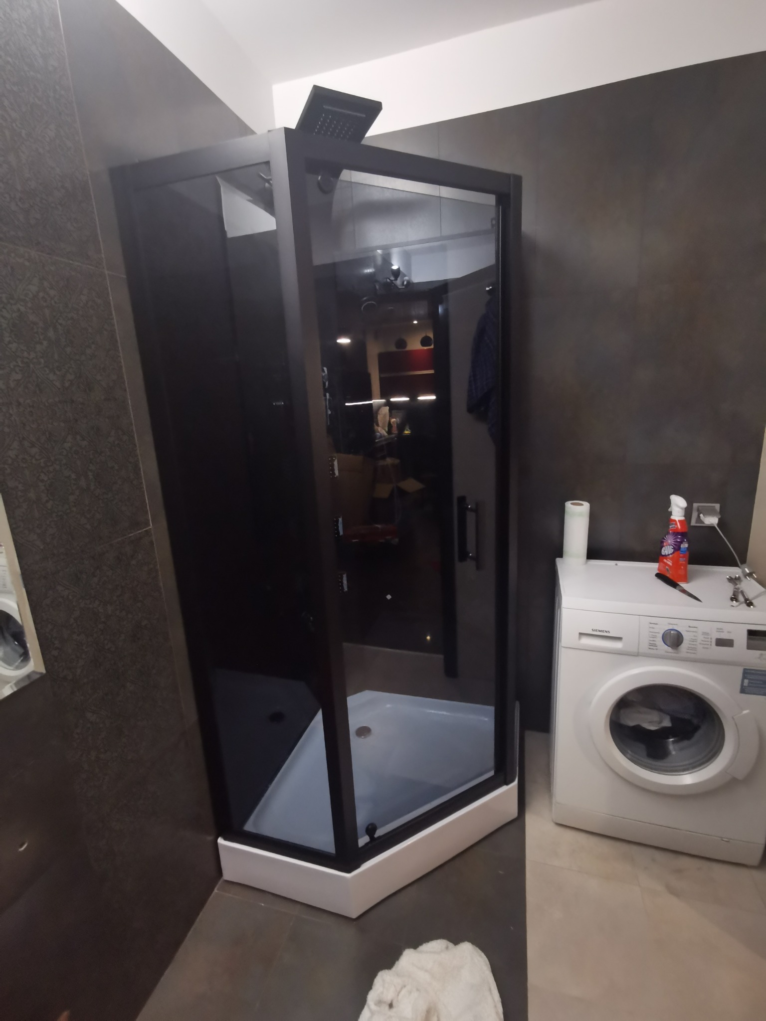 Czarna kabina prysznicowa z deszczownicą w narożniku łazienki, obok pralka i akcesoria czyszczące.