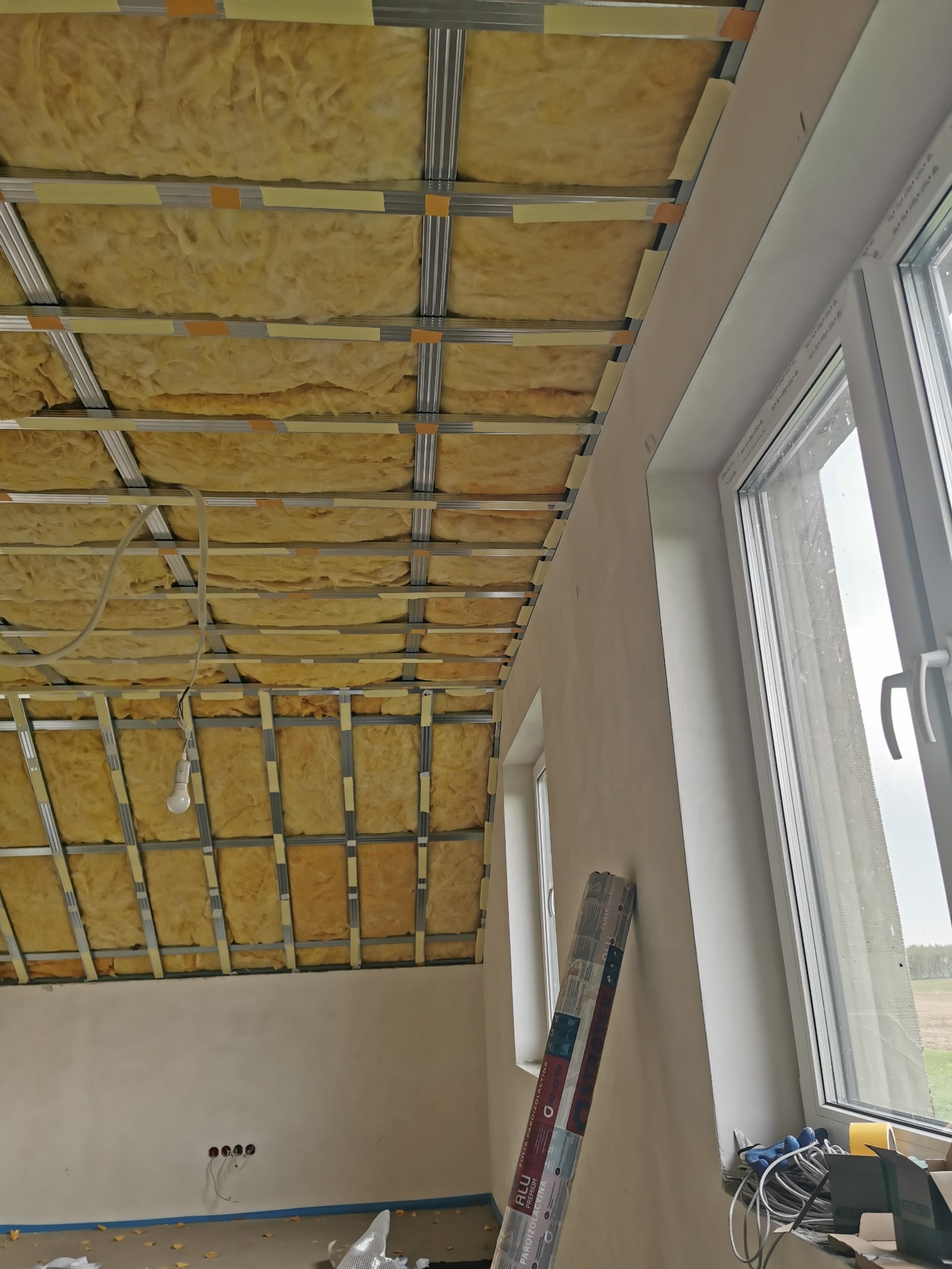 Montaż izolacji z wełny mineralnej w ruszcie sufitu podwieszanego z płyt karton-gips, widoczne okno i rolka folii paroizolacyjnej przy ścianie.