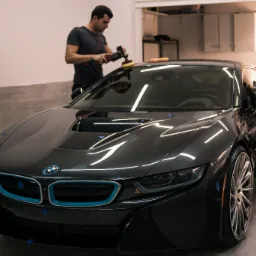 Polerowanie lakieru czarnego BMW i8 w studio detailingowym, widoczne niebieskie elementy karoserii zabezpieczone taśmą, pracownik używa polerki rotacyjnej, drugi pracownik wyciera karoserię...