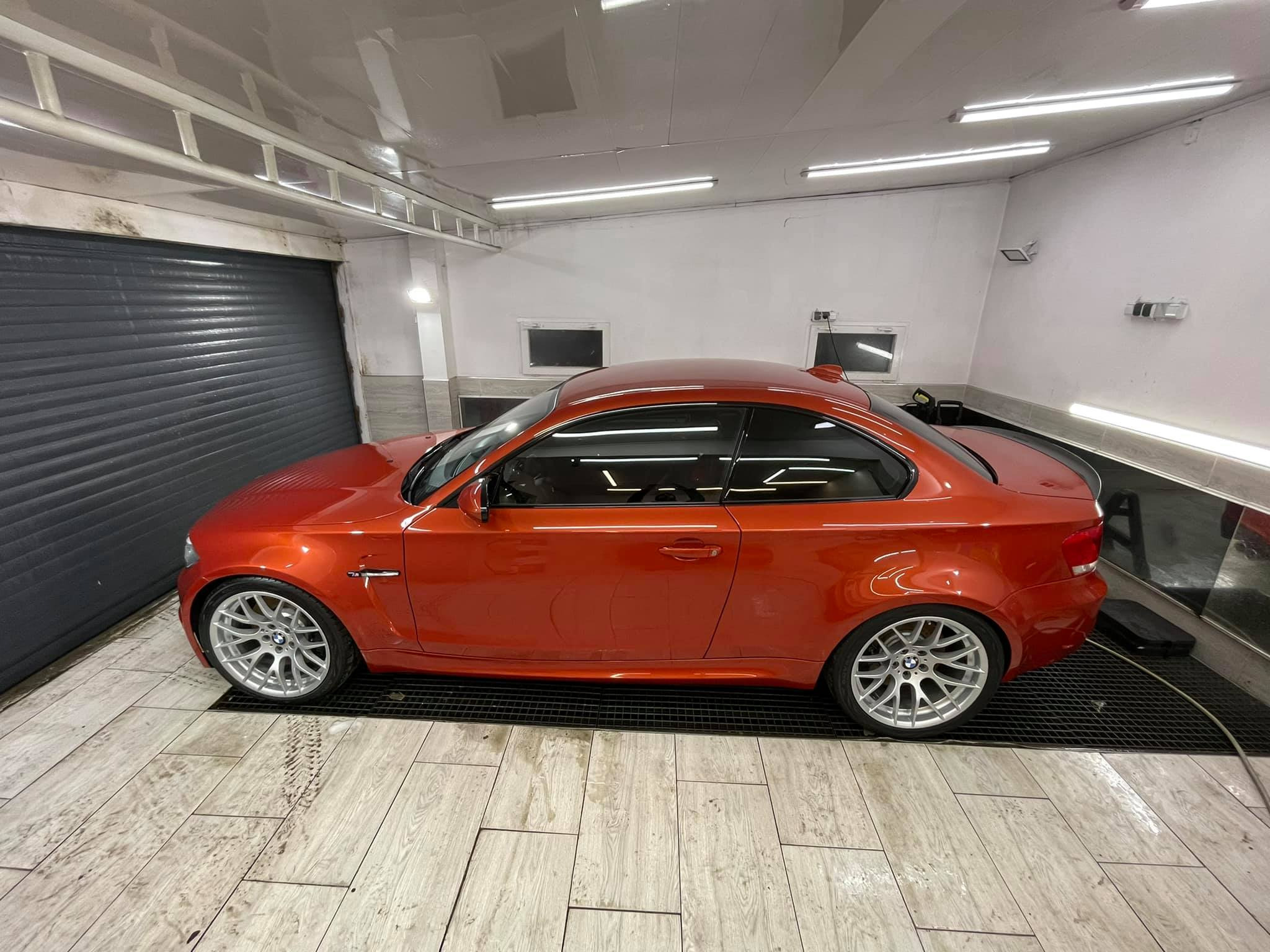 Pomarańczowe BMW serii 1 Coupe po umyciu, stojące w jasnym pomieszczeniu z szarą bramą rolowaną w tle.