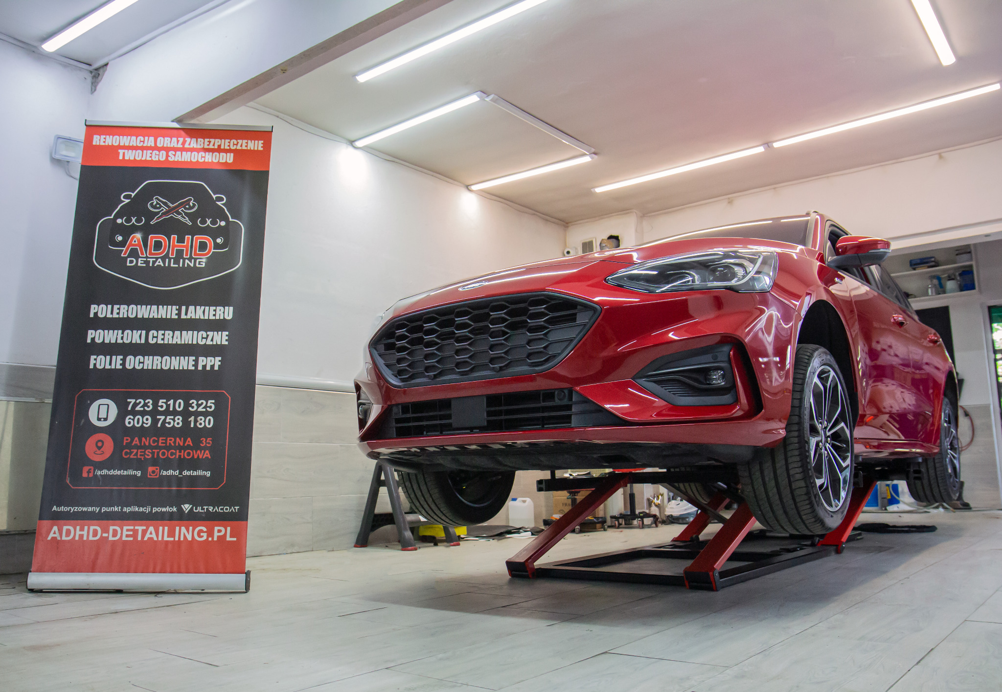 Czerwony samochód na podnośniku w warsztacie detailingowym, widoczny baner reklamowy ADHD Detailing z ofertą polerowania lakieru, powłok ceramicznych i folii ochronnych PPF.