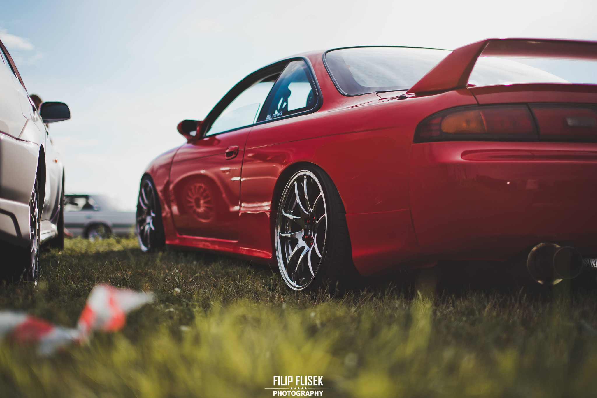 Czerwone sportowe auto z tuningowanymi felgami na trawie, widoczny podwójny wydech i spoiler, inne auta w tle.