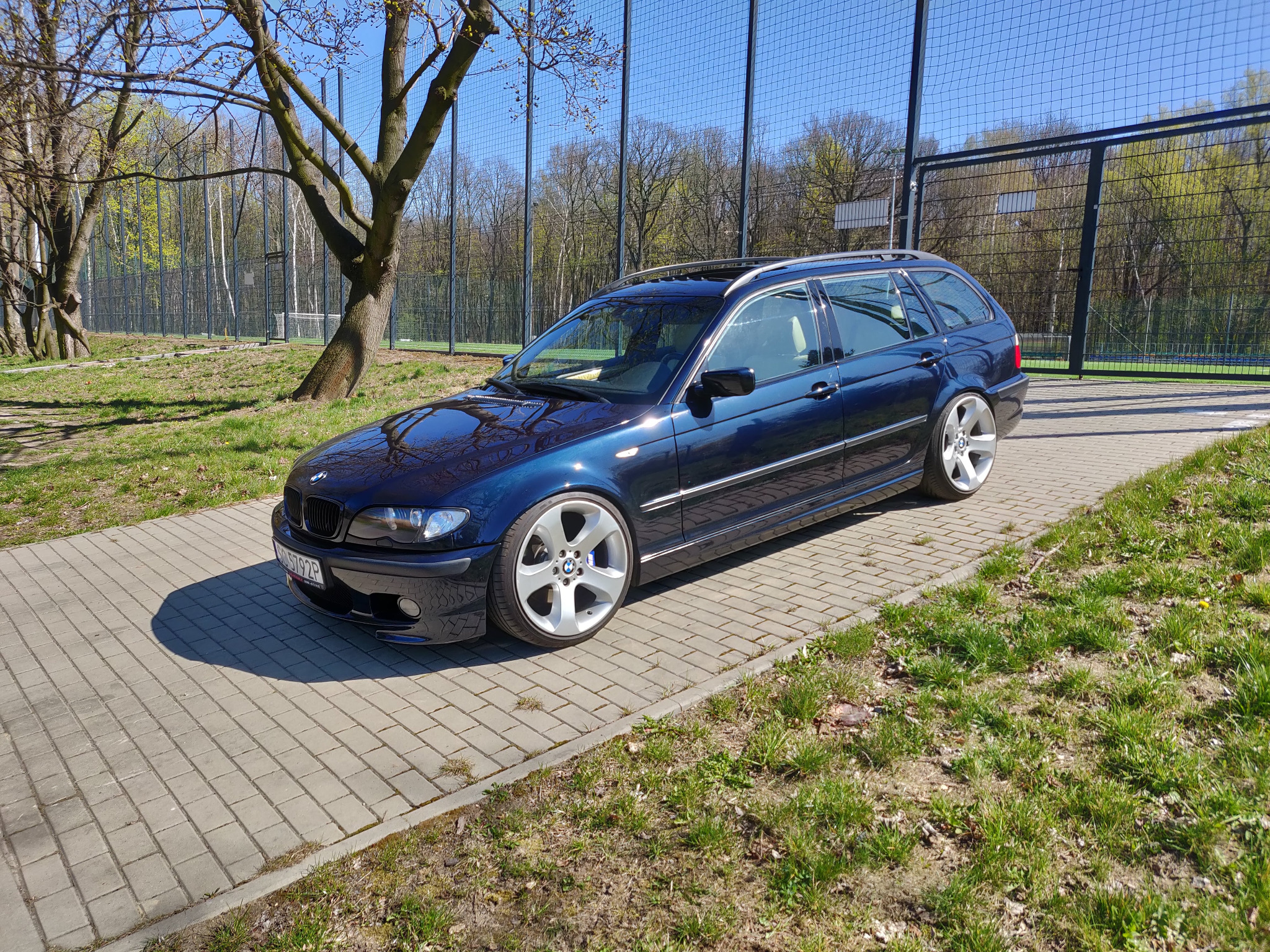 Granatowy samochód kombi marki BMW z felgami aluminiowymi stoi zaparkowany na brukowanym podjeździe w słoneczny dzień, z widoczną siatką ogrodzeniową i drzewami w tle.