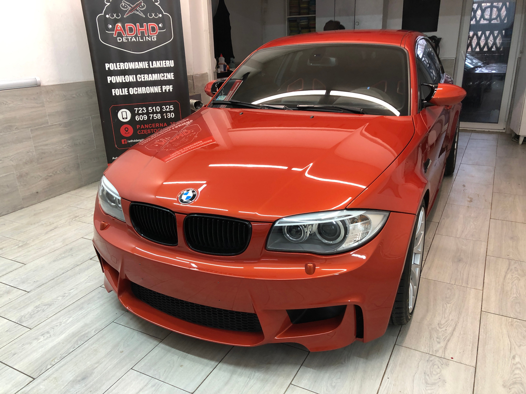 Błyszczący, pomarańczowy samochód BMW po detailingu, widoczne odbicia światła na lakierze, na tle ściany z logo firmy detailingowej 'ADHD Detailing'.