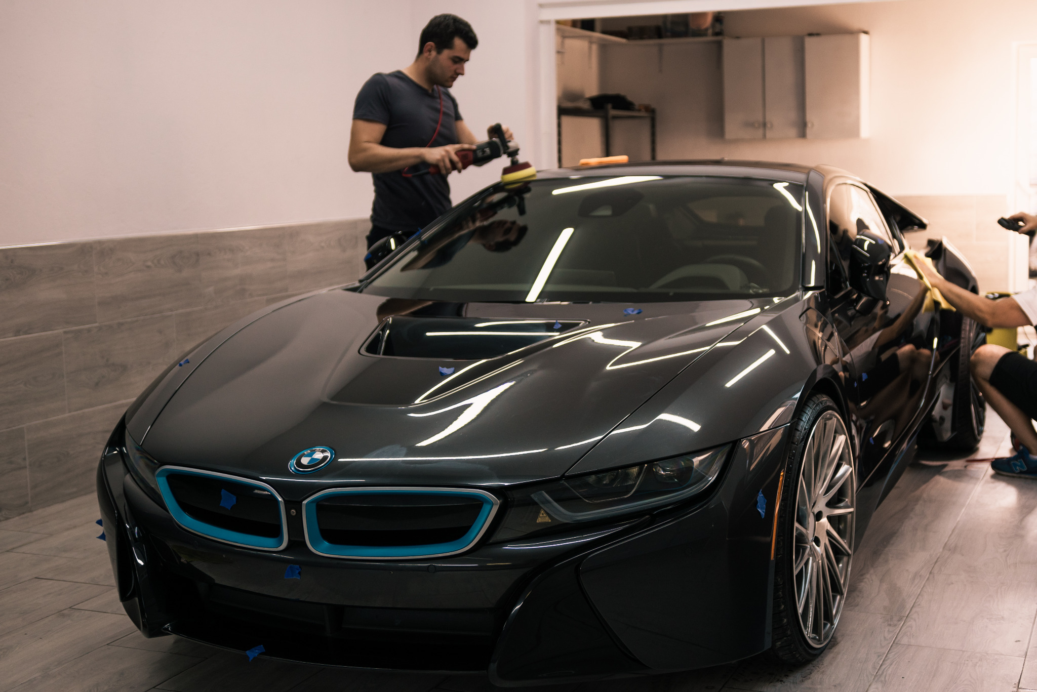 Polerowanie lakieru czarnego BMW i8 w studio detailingowym, widoczne niebieskie elementy karoserii zabezpieczone taśmą, pracownik używa polerki rotacyjnej, drugi pracownik wyciera karoserię...