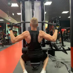 Mężczyzna ćwiczy na maszynie do wyciągania drążka w dół na siłowni, widok z tyłu, czarny strój sportowy, białe buty sportowe, żółto-czarne rękawice, w tle inne maszyny i osoby.