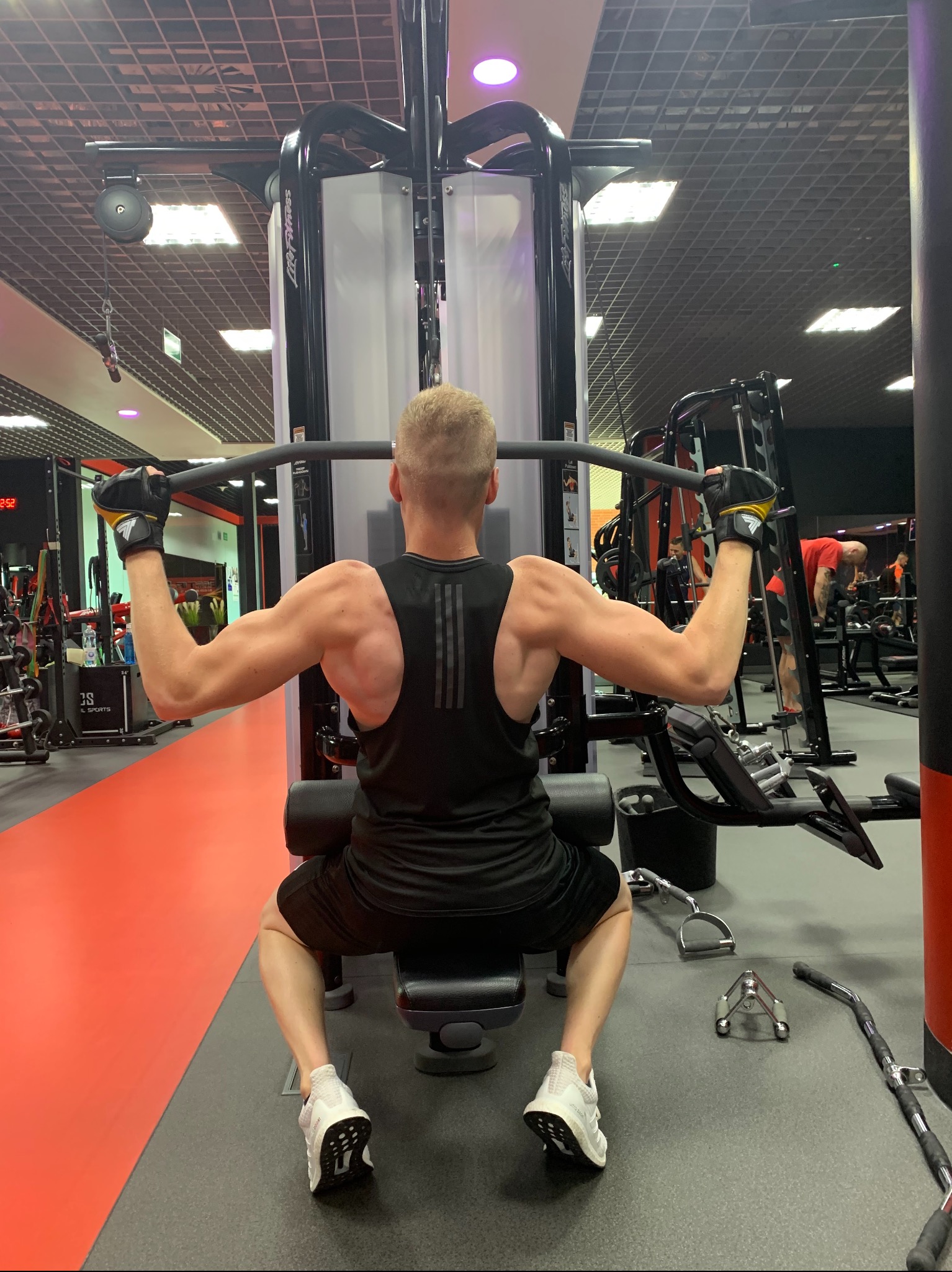 Mężczyzna ćwiczy na maszynie do wyciągania drążka w dół na siłowni, widok z tyłu, czarny strój sportowy, białe buty sportowe, żółto-czarne rękawice, w tle inne maszyny i osoby.