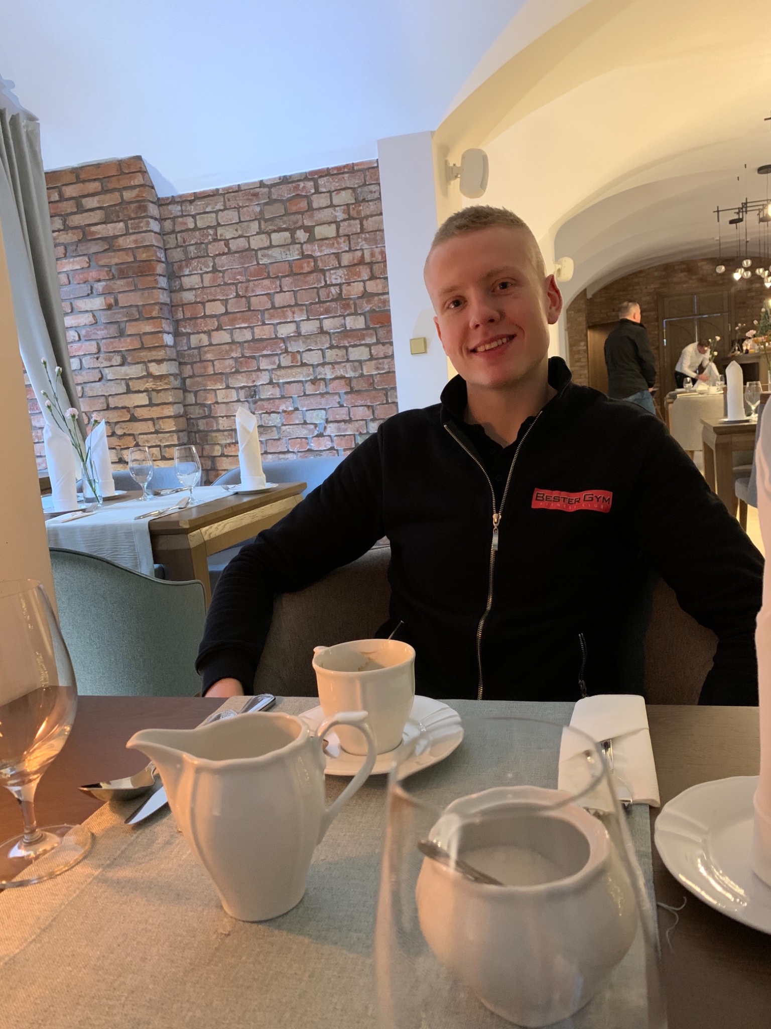 Uśmiechnięty mężczyzna w czarnej bluzie z logo 'Bester Gym' siedzi przy nakrytym stole z zastawą kawową w restauracji. W tle ceglana ściana i inni goście.