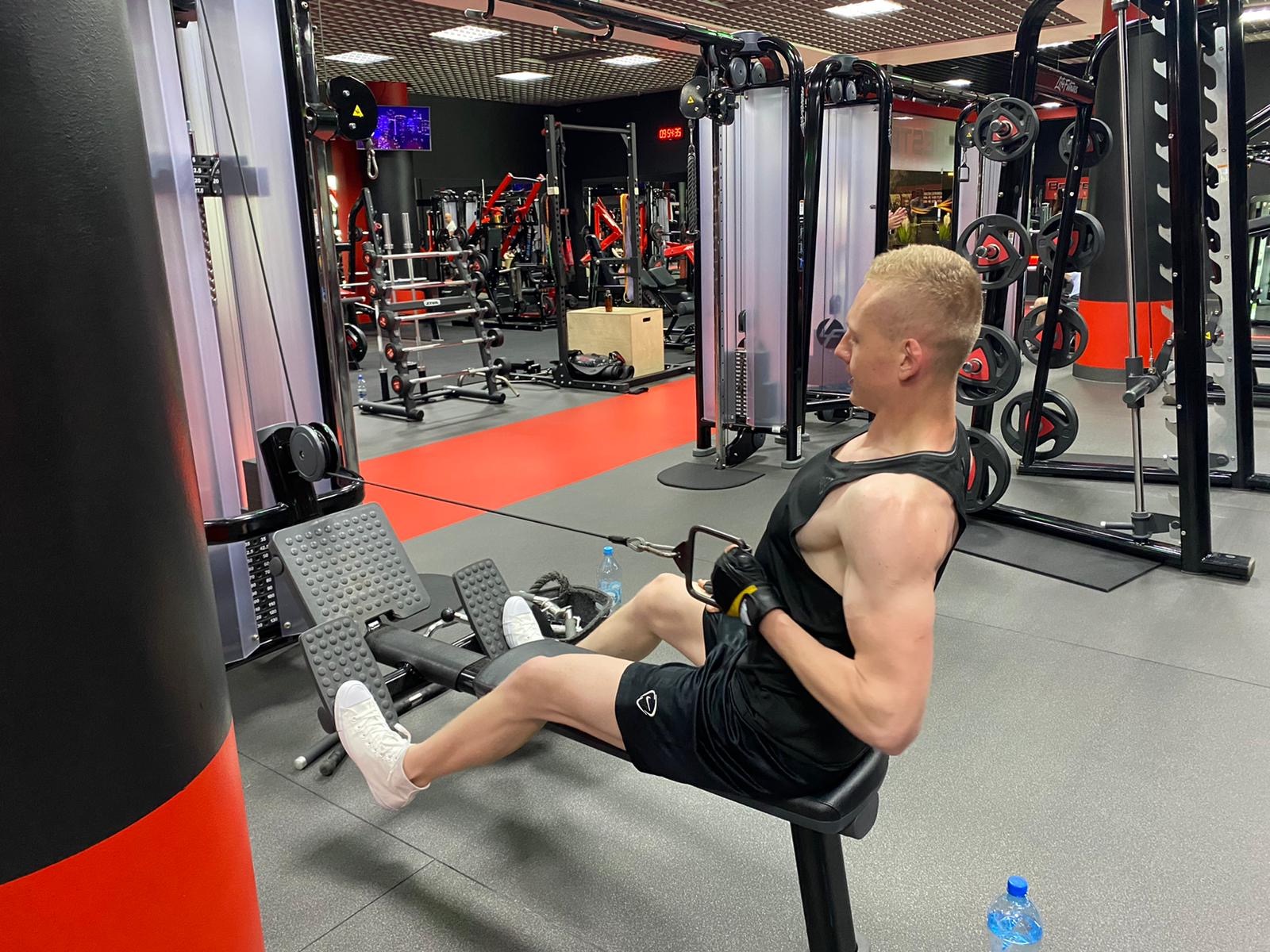 Mężczyzna w czarnym stroju sportowym wykonuje ćwiczenie na maszynie do wiosłowania w siłowni, widoczny profil twarzy, butelka wody obok.