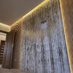 Nowoczesne wykończenie ściany z lameli i dekoracyjnej farby w odcieniach bieli i złota. Oświetlenie LED podkreśla strukturę. Widoczny fragment drzwi i podłogi.