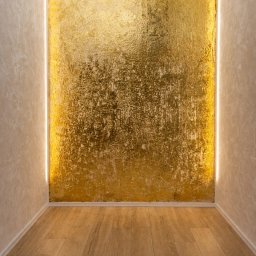 Bartosz Czynszak - Pokój z drewnianą podłogą i ścianą pokrytą złotą, teksturowaną farbą, oświetloną pionowymi pasami LED po bokach. Nowoczesny design.