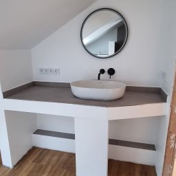 Bartosz Czynszak - Nowoczesna łazienka na poddaszu z owalną umywalką, okrągłym lustrem i drewnianą podłogą. Minimalistyczny design z czarną armaturą i szarą zabudową.