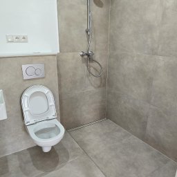 Bartosz Czynszak - Wykończona łazienka z szarymi płytkami, widoczna toaleta, prysznic i akcesoria. Na podłodze leżą tuby z silikonem. Minimalistyczny design.