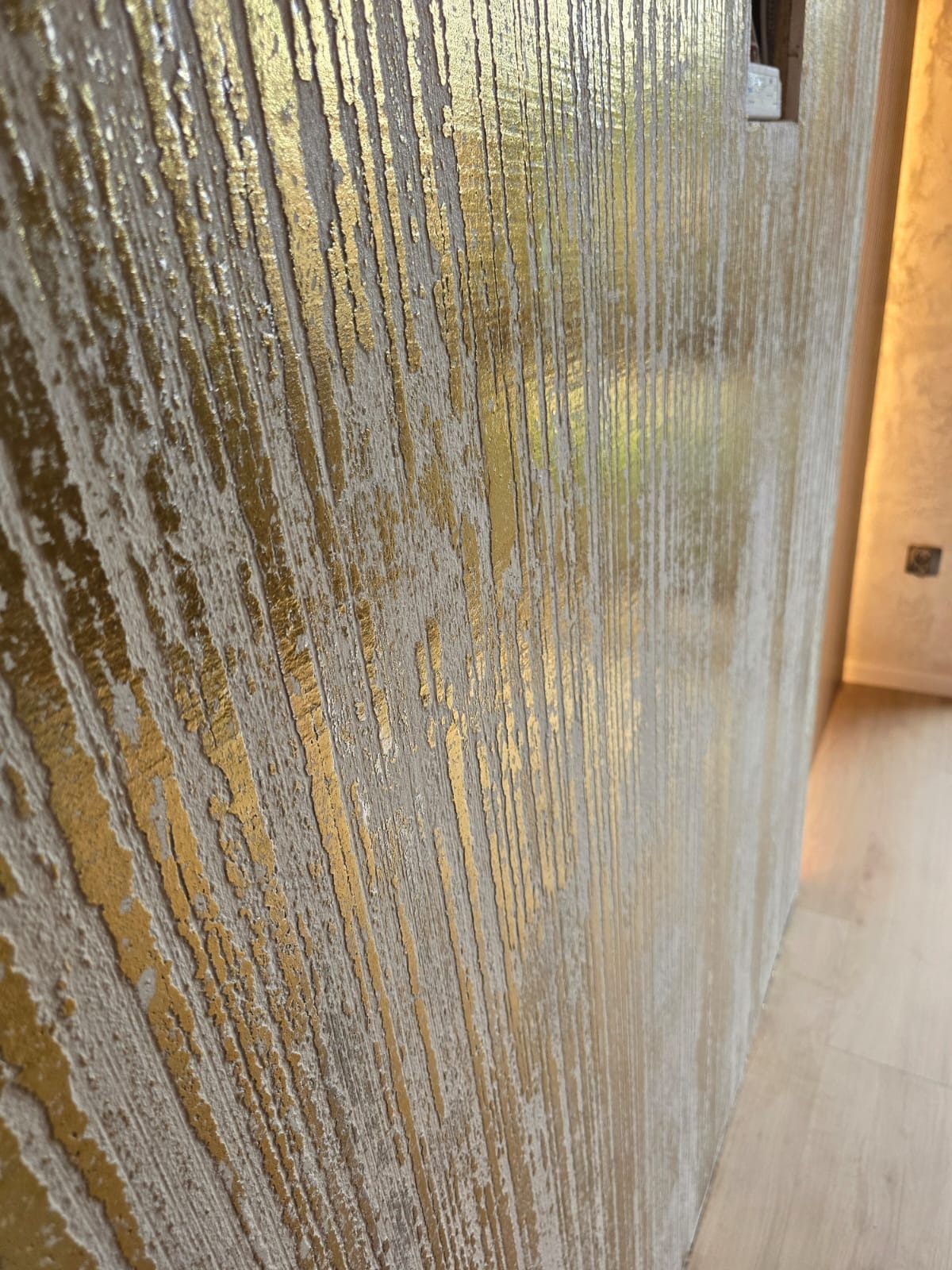 Dekoracyjna ściana z efektem złoconych, pionowych prążków na jasnym tle. Tekstura ściany jest wyraźnie widoczna, a całość prezentuje się elegancko i nowocześnie.