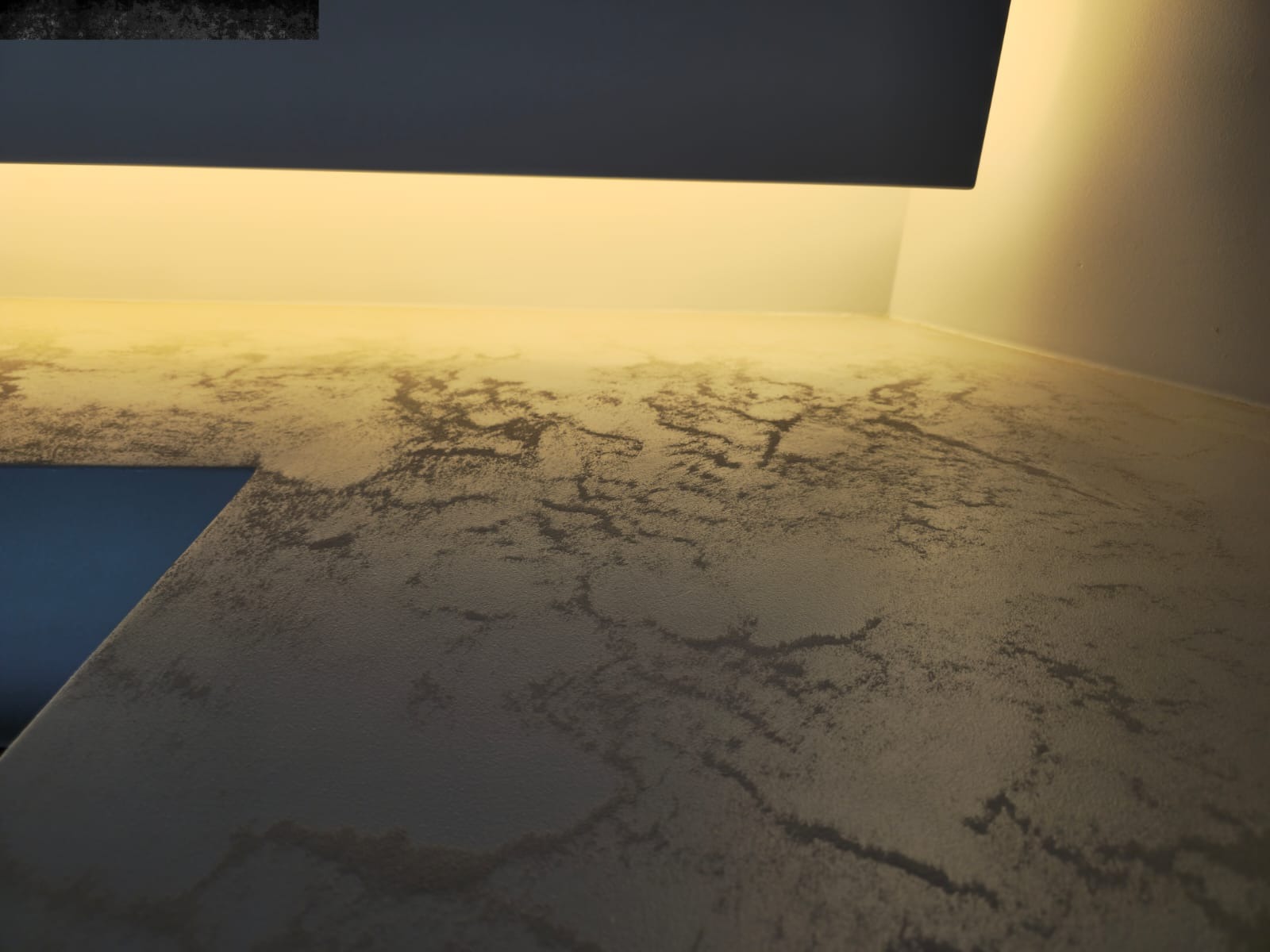 Beżowa ściana z dekoracyjną fakturą podświetlona ciepłym światłem, fragment blatu widoczny w lewym dolnym rogu kadru. Efektowna, nowoczesna gładź w pomieszczeniu.