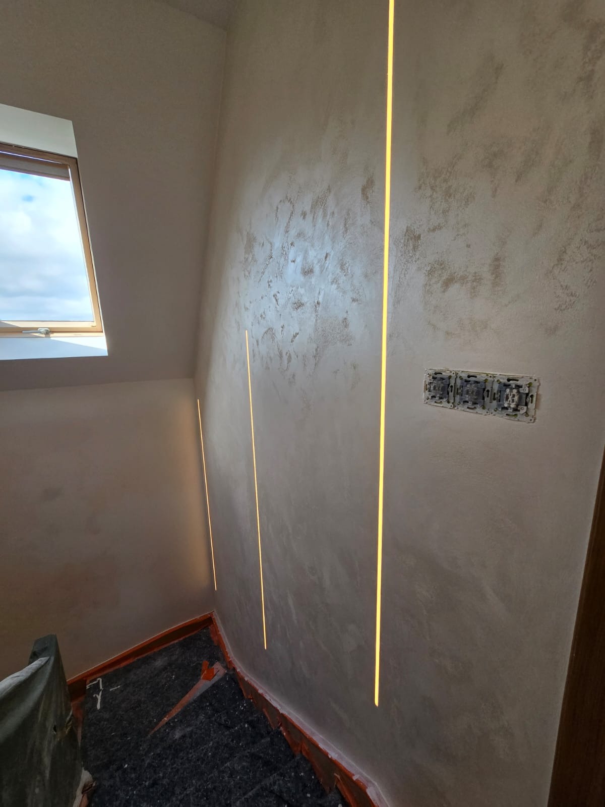 Ściana po gładzi dekoracyjnej z pionowymi pasami świetlnymi LED, widoczne gniazdka elektryczne i fragment schodów z zabezpieczeniem. Okno dachowe w tle.