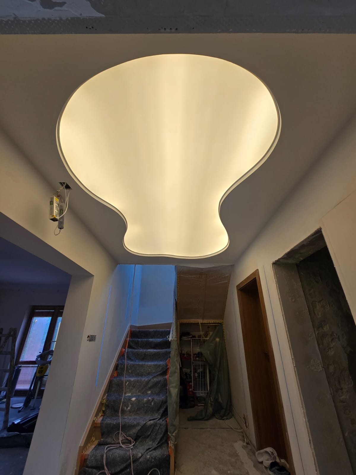 Nowoczesna lampa sufitowa o organicznym kształcie w korytarzu w trakcie wykańczania. Schody zabezpieczone folią, widoczne przygotowania do prac wykończeniowych.
