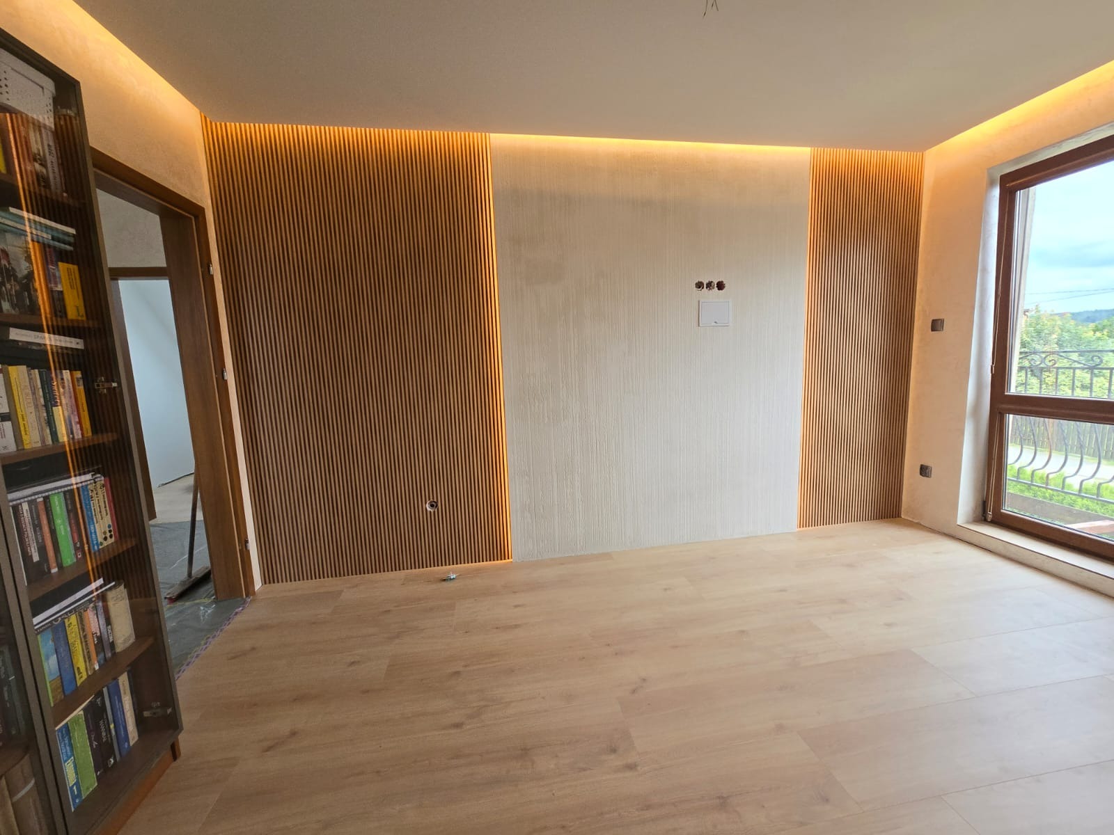 Nowoczesne wykończenie wnętrza z drewnianymi lamelami ściennymi i oświetleniem LED, jasna podłoga, widok na zewnątrz przez okno. Minimalistyczny design.