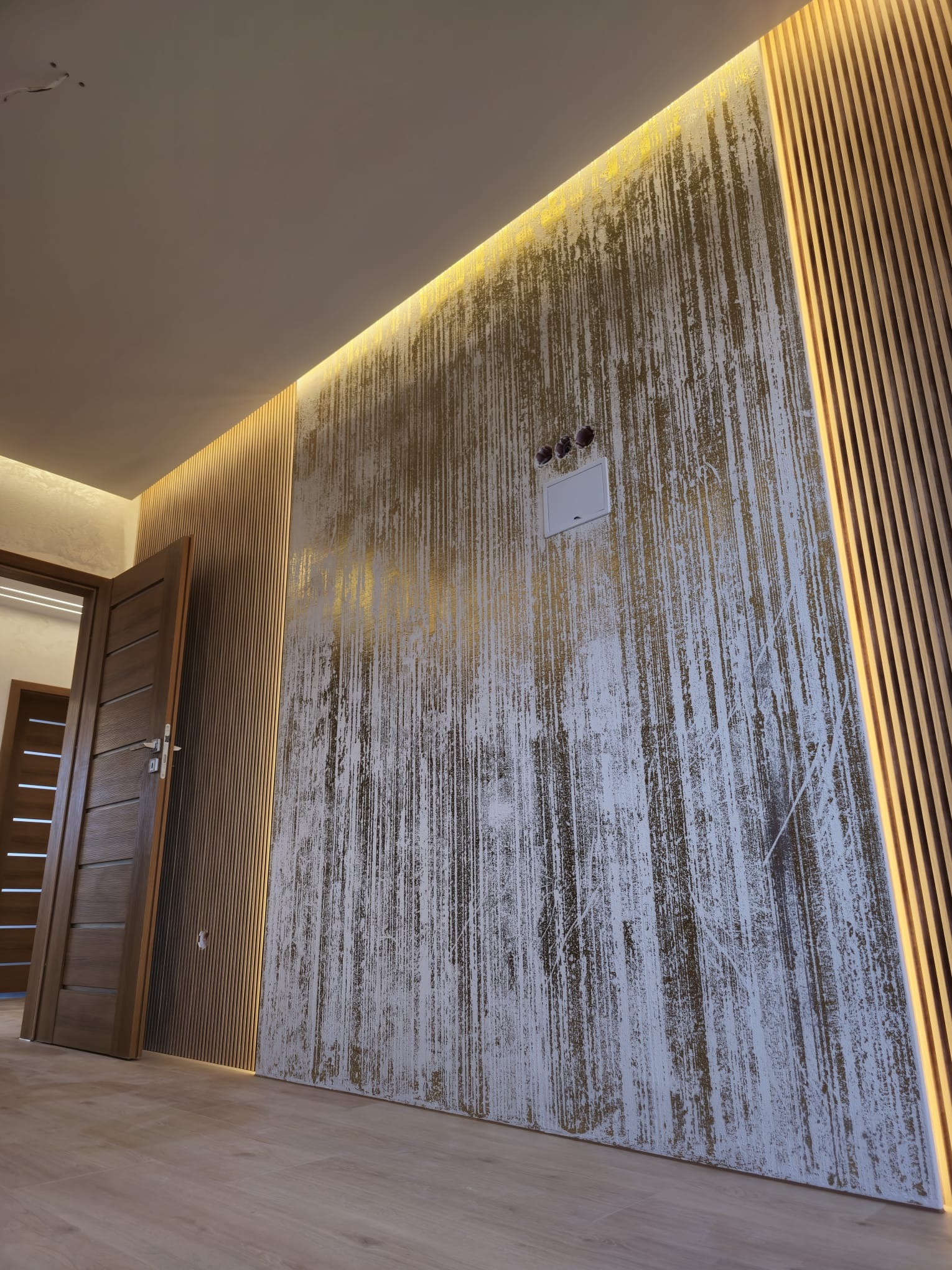 Nowoczesne wykończenie ściany z lameli i dekoracyjnej farby w odcieniach bieli i złota. Oświetlenie LED podkreśla strukturę. Widoczny fragment drzwi i podłogi.