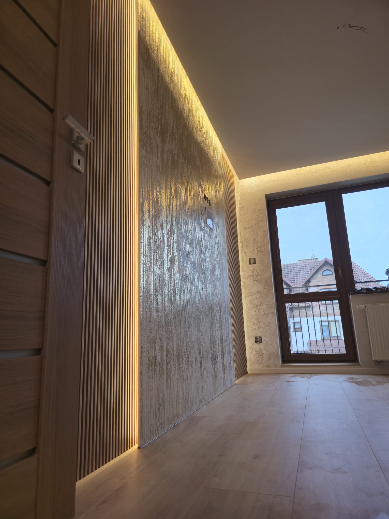 Wykończone wnętrze: drewniane panele ścienne z oświetleniem LED, dekoracyjna ściana w odcieniach beżu, drewniana podłoga i okno z widokiem na dom.