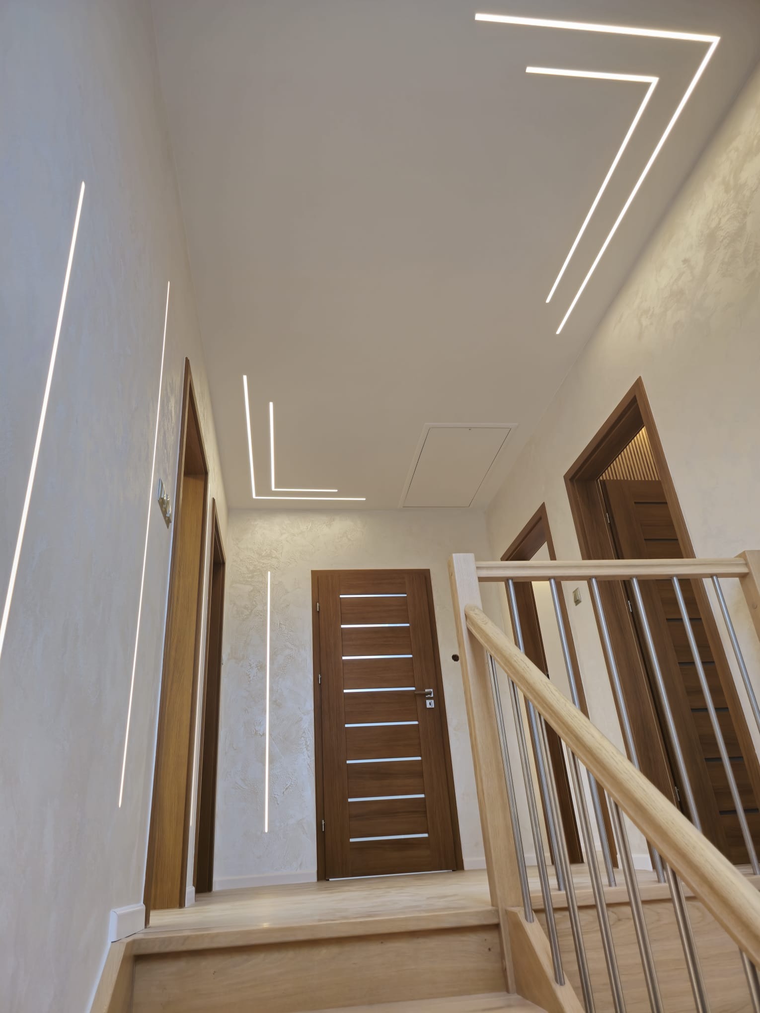 Nowoczesne oświetlenie LED w korytarzu z drewnianymi drzwiami i schodami. Minimalistyczny design z liniowymi lampami na suficie i ścianach.