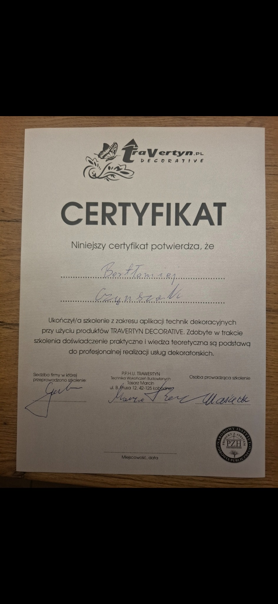 Certyfikat ukończenia szkolenia z technik dekoracyjnych Travertyn Decorative, z podpisami i pieczęcią PZH, leżący na drewnianym podłożu. Dokument potwierdza kwalifikacje.