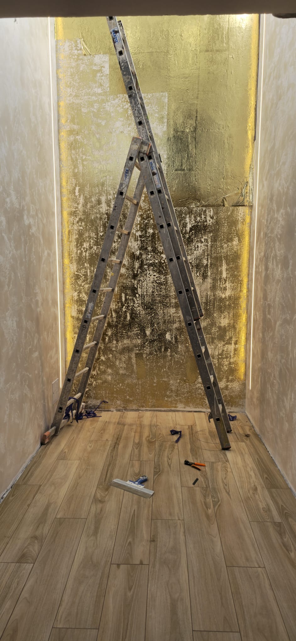 Pokój w trakcie wykańczania: złota ściana z ledowym oświetleniem, drewnopodobna podłoga, drabina i narzędzia malarskie. Widok z dołu, podkreślający wysokość pomieszczenia.