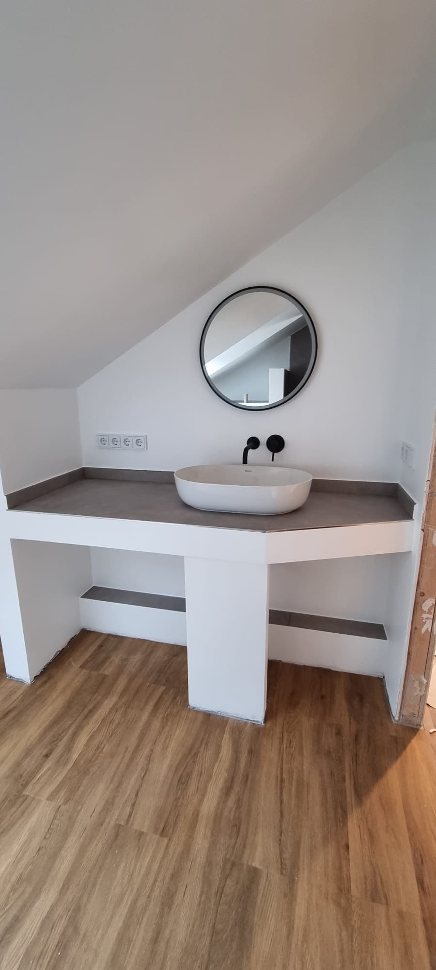 Nowoczesna łazienka na poddaszu z owalną umywalką, okrągłym lustrem i drewnianą podłogą. Minimalistyczny design z czarną armaturą i szarą zabudową.