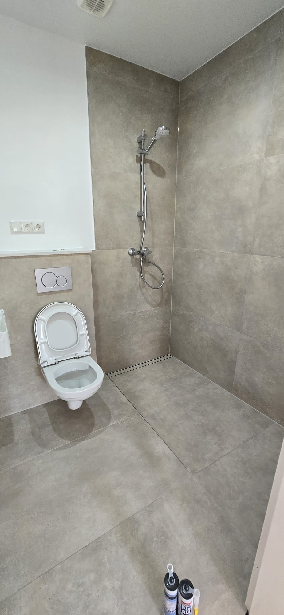 Wykończona łazienka z szarymi płytkami, widoczna toaleta, prysznic i akcesoria. Na podłodze leżą tuby z silikonem. Minimalistyczny design.