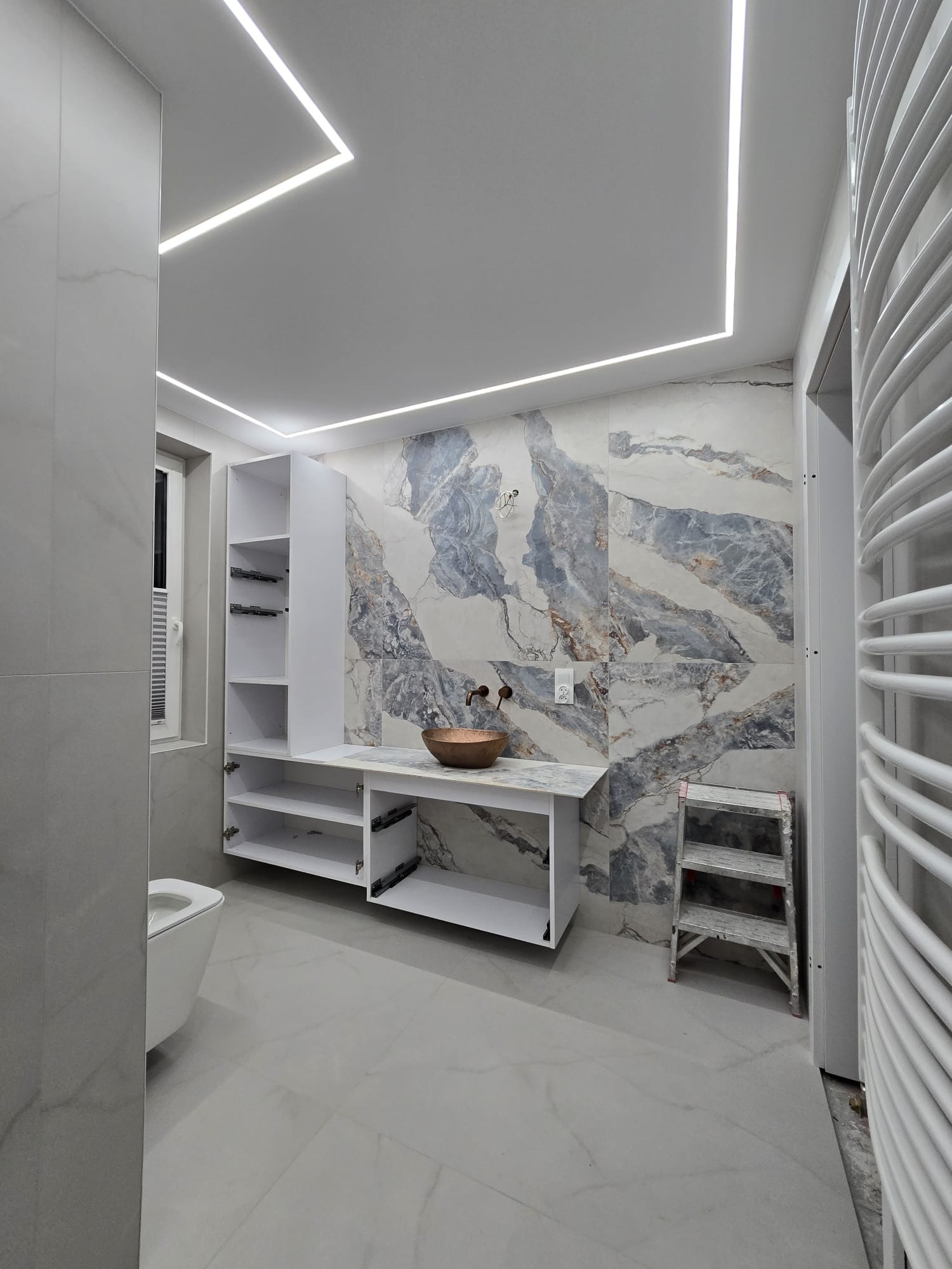 Nowoczesna łazienka z marmurowymi płytkami, podświetlanym sufitem LED i miedzianą umywalką. Widoczna biała zabudowa meblowa i drabinka.