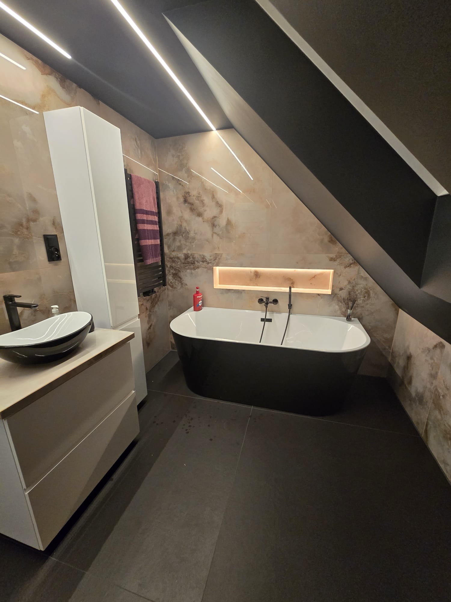 Nowoczesna łazienka na poddaszu z czarną wanną wolnostojącą, umywalką nablatową i marmurowymi płytkami. Oświetlenie LED podkreśla skośny sufit i dodaje elegancji.