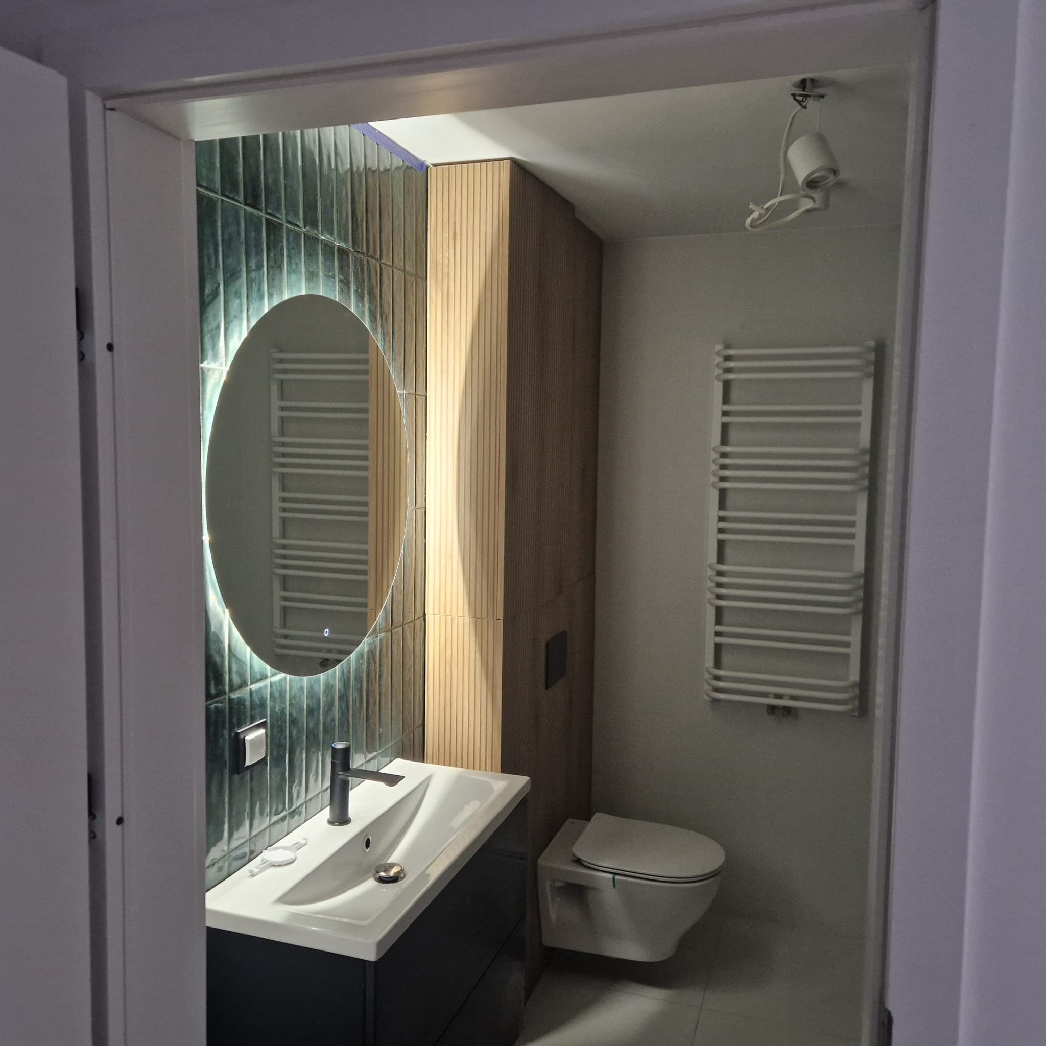 Nowoczesna łazienka po remoncie: zielone płytki, okrągłe lustro z podświetleniem LED, drewniana zabudowa, biała umywalka i toaleta podwieszana.