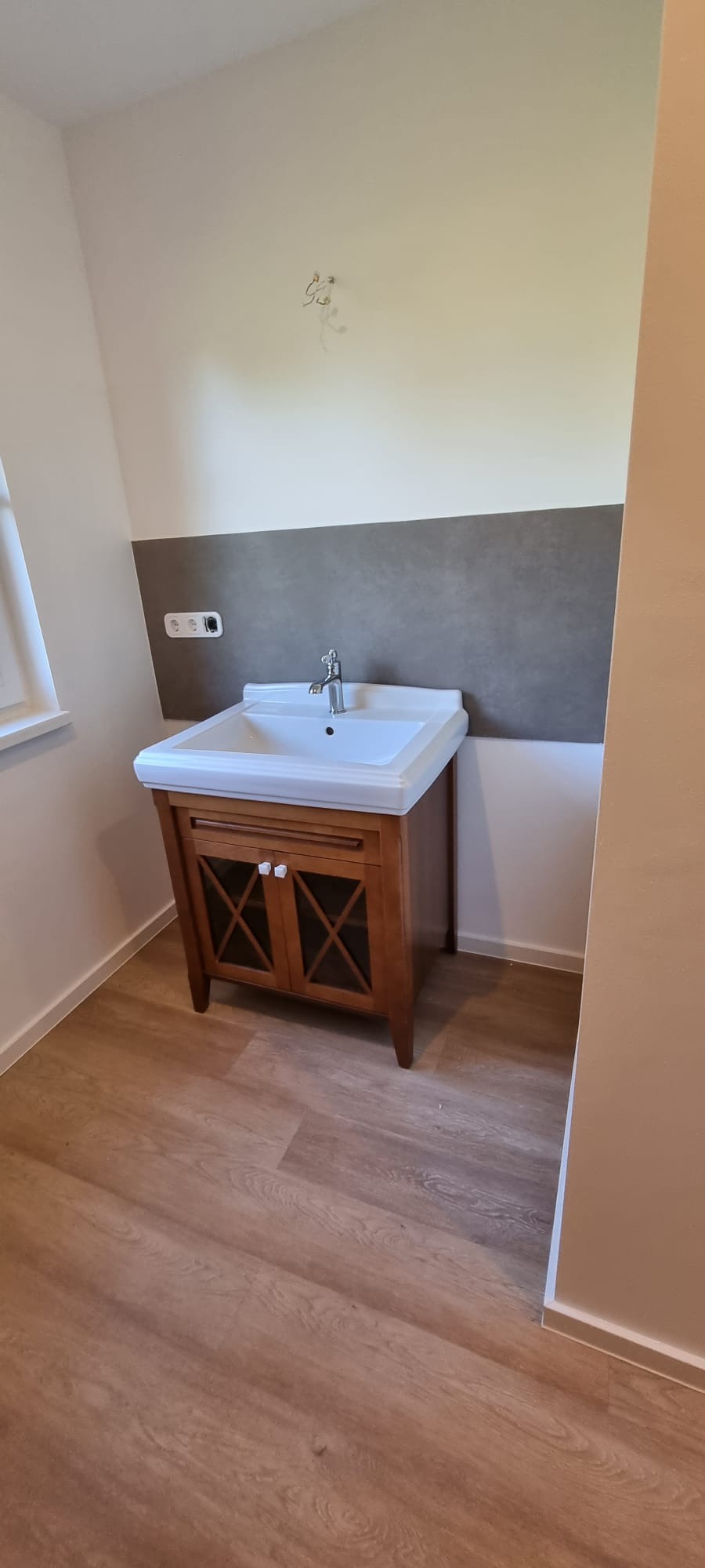 Wykończona łazienka z drewnianą szafką pod umywalką, szarą płytką na ścianie i panelami podłogowymi. Widoczne gniazdko elektryczne i niedokończone mocowanie lampy.