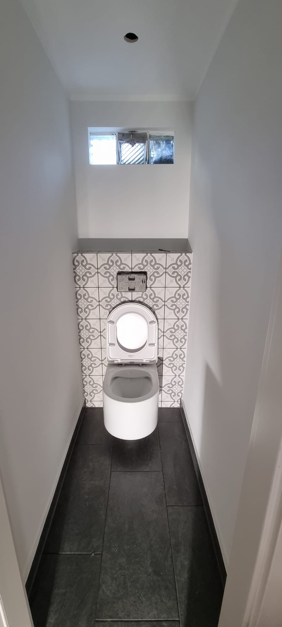 Kompaktowa toaleta w wąskiej łazience z czarną podłogą i dekoracyjnymi płytkami nad zabudową spłuczki. Małe okno z luksferami w górnej części ściany.