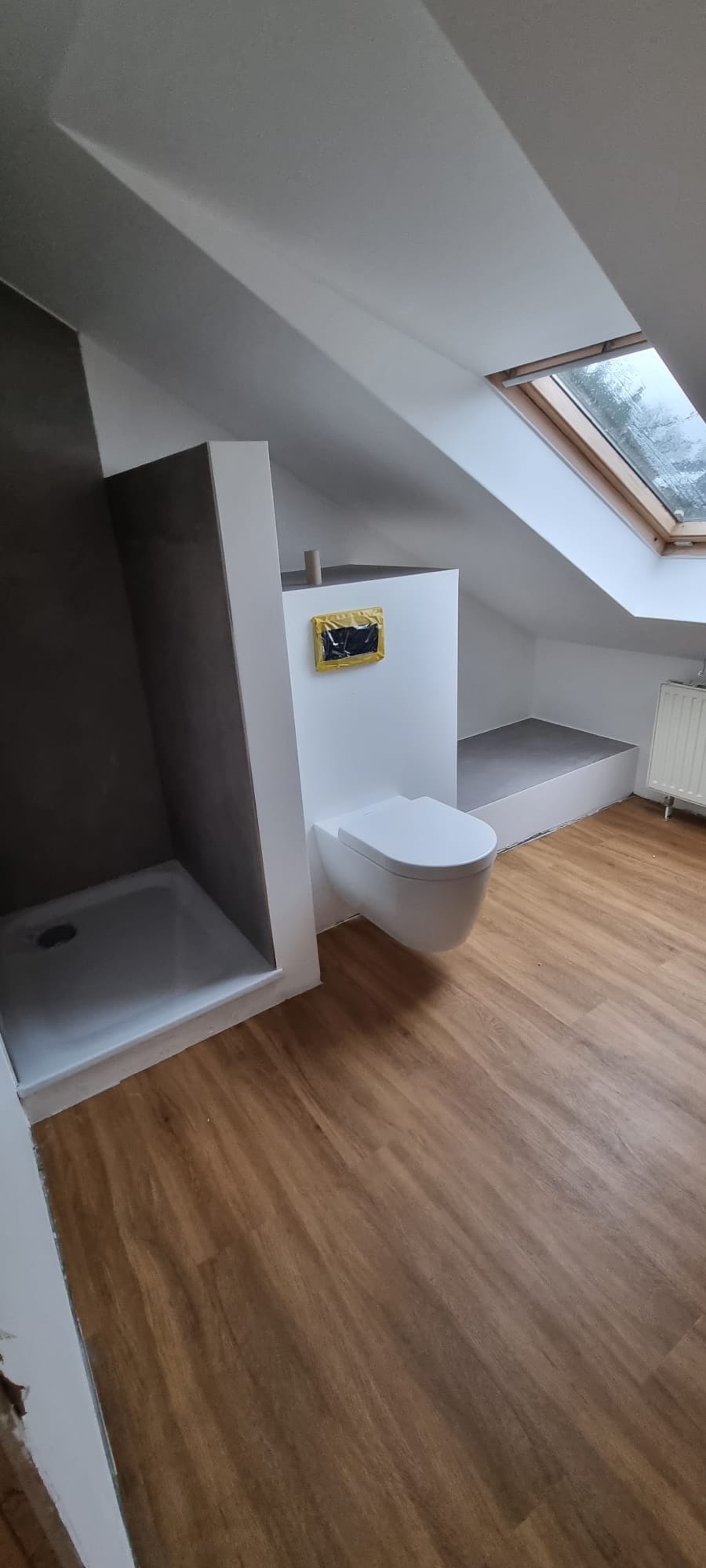 Nowoczesna łazienka na poddaszu z kabiną prysznicową typu walk-in, podwieszaną toaletą i drewnianą podłogą. Okno dachowe wpuszcza naturalne światło.