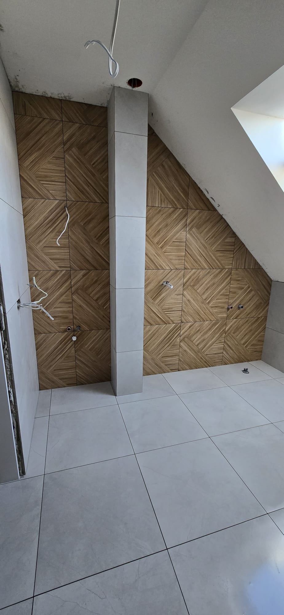 Wykończona łazienka na poddaszu z drewnopodobnymi płytkami ułożonymi w geometryczny wzór na ścianach i jasnymi płytkami na podłodze. Widoczne instalacje.