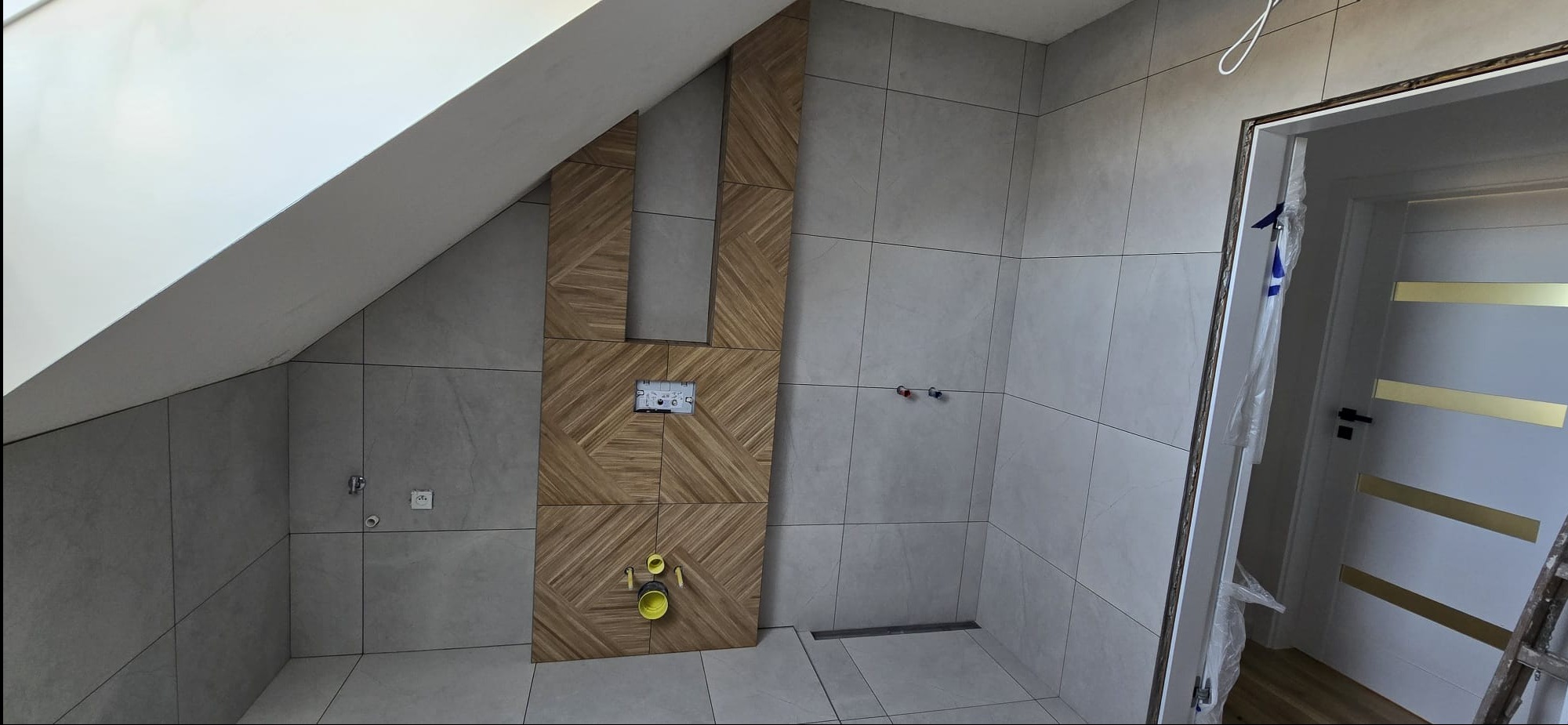 Wykończona łazienka na poddaszu z szarymi płytkami i dekoracyjną ścianą z drewnopodobnych płytek. Widoczna instalacja podtynkowa i odpływ liniowy.