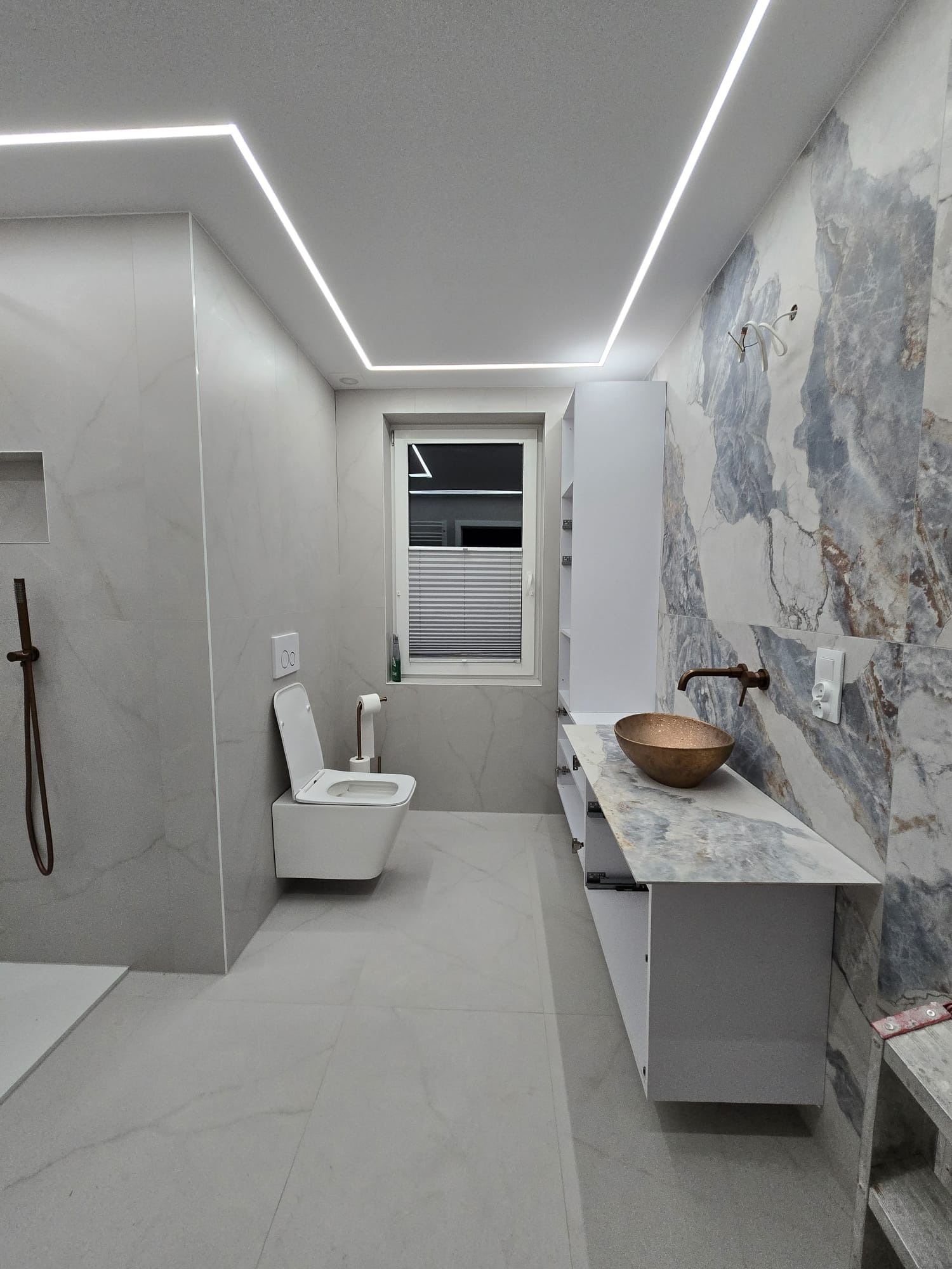 Nowoczesna łazienka z marmurowymi płytkami, podświetlanym sufitem LED, miedzianą umywalką i minimalistyczną toaletą. Widok na okno z żaluzjami.