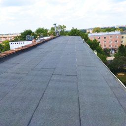 Dekadach - Płaski dach pokryty szarą papą termozgrzewalną na budynku wielorodzinnym, widok z góry na miasto w tle, widoczny fragment instalacji odgromowej.