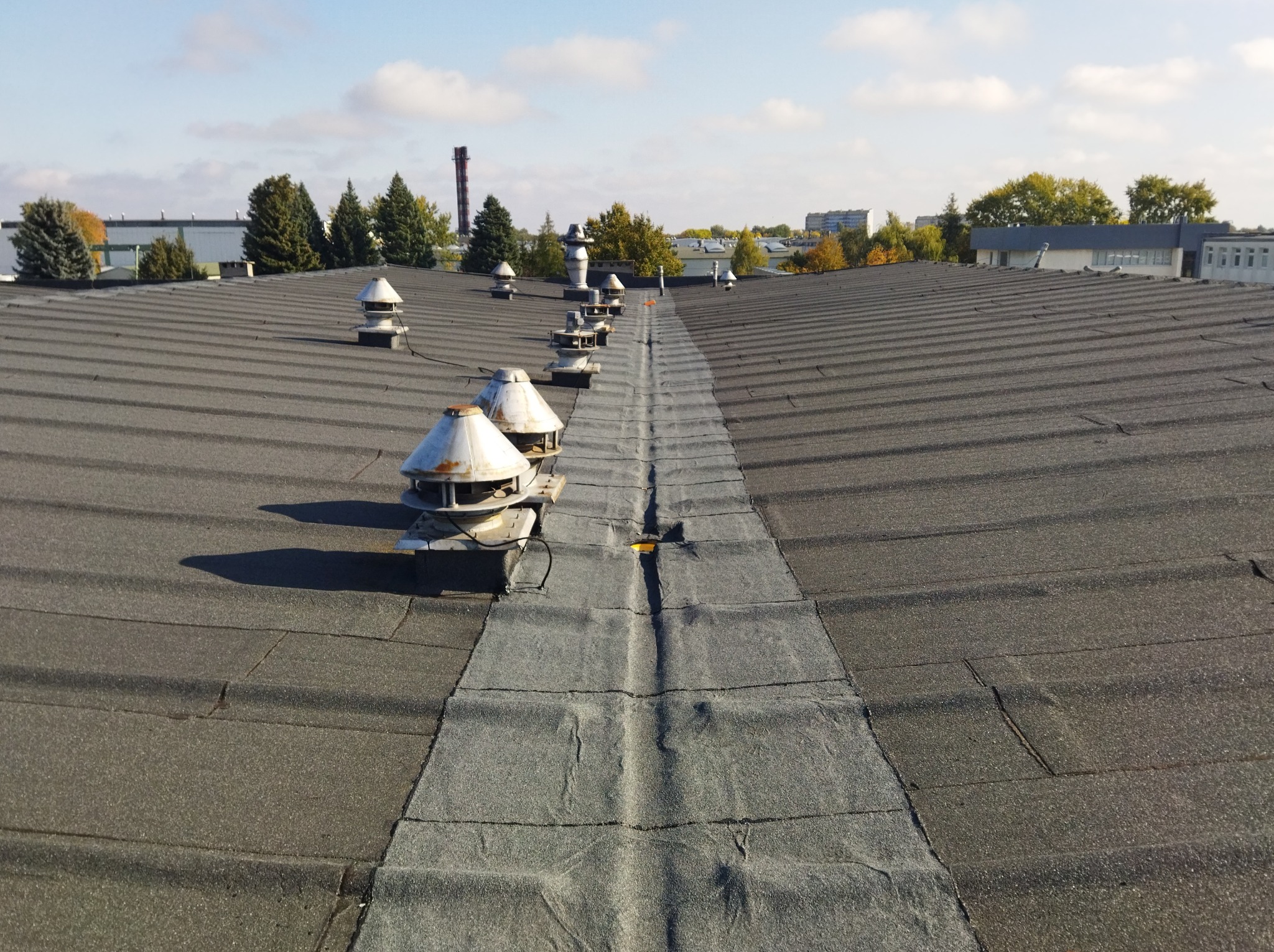 Płaski dach pokryty papą z kominkami wentylacyjnymi i pasem wzmocnienia na środku, widok z góry w słoneczny dzień.