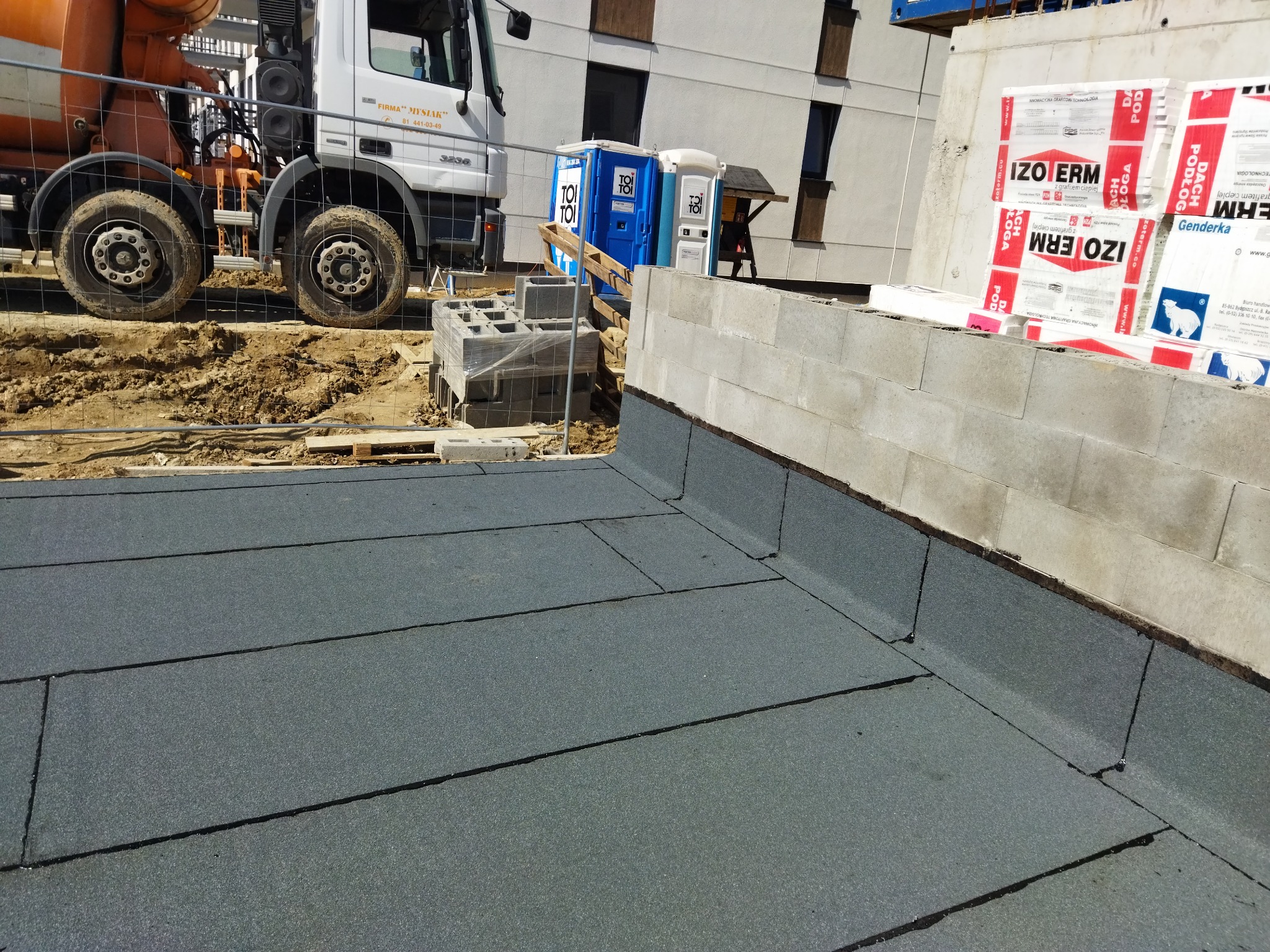 Szczegółowe ujęcie płaskiego dachu pokrytego papą termozgrzewalną na budowie, widoczny fragment ściany attyki, w tle betoniarka, przenośne toalety i budynek w trakcie budowy.