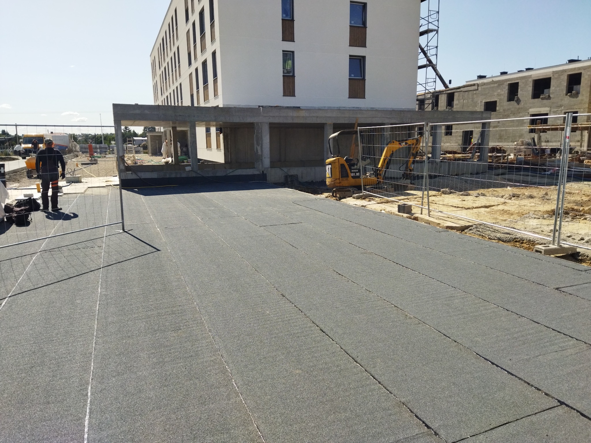 Przygotowanie podłoża pod przyszłe pokrycie dachu: rozłożona warstwa izolacyjna na budowie z widocznym mini-koparką CAT i pracownikiem w tle.