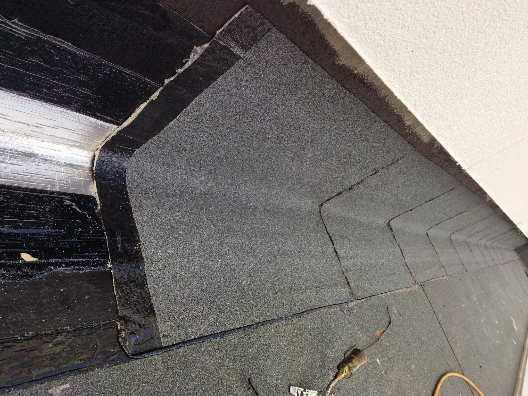 Fragment dachu z papy termozgrzewalnej, widoczne łączenia i narzędzie do zgrzewania, zarys białej ściany w tle.