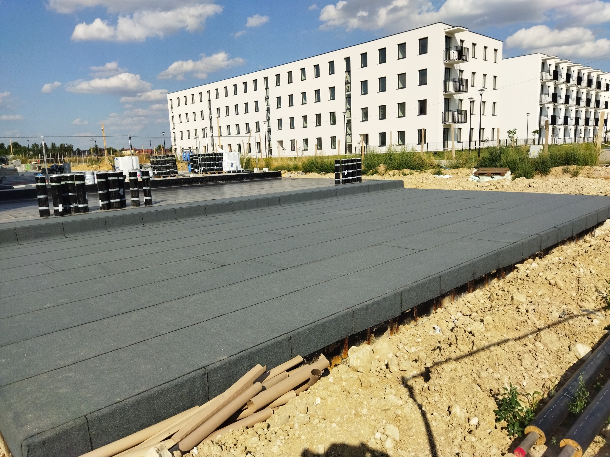 Płaski dach pokryty szarą membraną bitumiczną w trakcie realizacji, widoczne rolki materiału izolacyjnego i nowo wybudowany budynek mieszkalny w tle.